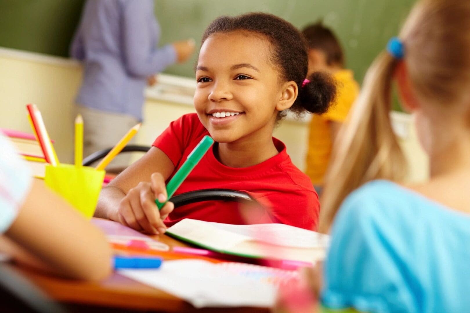 A young girl smiles while holding a green pencil.